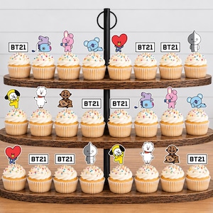 Peut inclure: Un pr&eacute;sentoir &agrave; cupcakes en bois &agrave; trois niveaux contenant vingt-quatre cupcakes garnis de vermicelles color&eacute;s. Chaque cupcake est surmont&eacute; d'une d&eacute;coration en papier repr&eacute;sentant divers personnages BT21 ou le texte BT21 dans un cadre noir.