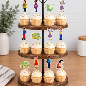 May include: A three-tiered wooden cupcake stand holding twelve cupcakes, each topped with a paper cutout of characters and a school bus from The Magic School Bus. The cupcakes feature white frosting with colorful sprinkles in paper liners.