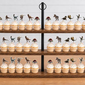 Pu&ograve; includere: Un'alzata per cupcake in legno a tre livelli che ospita numerosi cupcake sormontati da varie figure di dinosauri e decorazioni circolari con il logo di Jurassic Park. Il supporto presenta un'asta centrale in metallo nero.
