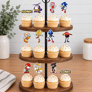 May include: A three-tiered wooden cupcake stand holding twelve cupcakes topped with white frosting and colorful sprinkles. Each cupcake features a paper topper depicting various Sonic the Hedgehog characters or the Sonic logo on a wooden pick.