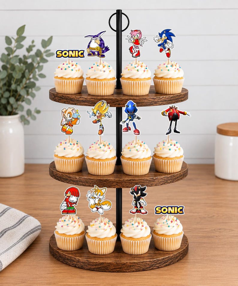 May include: A three-tiered wooden cupcake stand holding twelve cupcakes topped with white frosting and colorful sprinkles. Each cupcake features a paper topper depicting various Sonic the Hedgehog characters or the Sonic logo on a wooden pick.