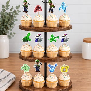 Puede incluir: Un soporte de madera para cupcakes de tres niveles con doce cupcakes cubiertos de glaseado blanco y chispas de colores. Cada cupcake tiene un adorno de papel decorativo que representa personajes de la serie de juegos Luigi's Mansion.