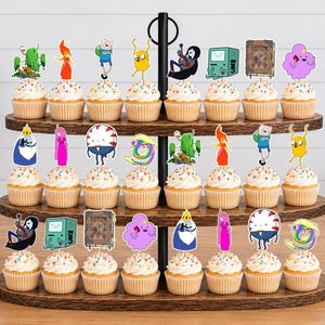 K&ouml;nnte beinhalten: Ein dreist&ouml;ckiger Cupcake-St&auml;nder aus Holz mit vierundzwanzig Cupcakes, die mit bunten Papieraufsteckern verziert sind, welche verschiedene Adventure Time-Figuren wie Finn, Jake, Prinzessin Bubblegum und den Eisk&ouml;nig zeigen.