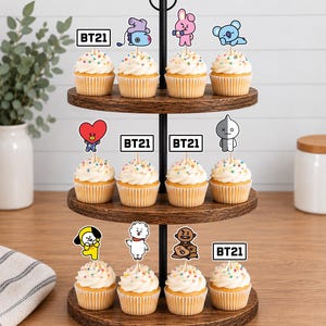 Peut inclure: Un pr&eacute;sentoir &agrave; cupcakes en bois &agrave; trois niveaux contenant douze cupcakes garnis de gla&ccedil;age blanc, de vermicelles color&eacute;s et de d&eacute;corations en papier repr&eacute;sentant les personnages BT21 avec le texte BT21 sur un support en m&eacute;tal noir.