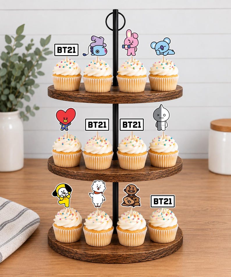 Peut inclure: Un pr&eacute;sentoir &agrave; cupcakes en bois &agrave; trois niveaux contenant douze cupcakes garnis de gla&ccedil;age blanc, de vermicelles color&eacute;s et de d&eacute;corations en papier repr&eacute;sentant les personnages BT21 avec le texte BT21 sur un support en m&eacute;tal noir.