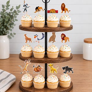 K&ouml;nnte beinhalten: Ein dreist&ouml;ckiger Cupcake-St&auml;nder aus Holz mit zw&ouml;lf Cupcakes, die mit wei&szlig;em Zuckerguss und bunten Streuseln verziert sind. Jeder Cupcake ist mit einem Papieraufsteller eines Charakters aus Der K&ouml;nig der L&ouml;wen versehen, darunter Simba, Mufasa, Scar, Timon, Pumbaa und Zazu.