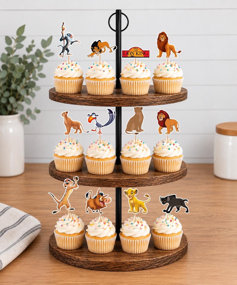 K&ouml;nnte beinhalten: Ein dreist&ouml;ckiger Cupcake-St&auml;nder aus Holz mit zw&ouml;lf Cupcakes, die mit wei&szlig;em Zuckerguss und bunten Streuseln verziert sind. Jeder Cupcake ist mit einem Papieraufsteller eines Charakters aus Der K&ouml;nig der L&ouml;wen versehen, darunter Simba, Mufasa, Scar, Timon, Pumbaa und Zazu.