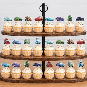 May include: A three-tiered wooden cupcake stand holding twenty-three cupcakes, each topped with a colorful paper cutout of a character from the Cars movie franchise, featuring various vehicle designs in red, blue, green, and purple hues.