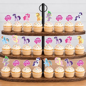 Op de afbeelding: Een houten cupcake-standaard met drie niveaus met vierentwintig cupcakes, versierd met kleurrijke My Little Pony-papieren toppers en My Little Pony-logo's. De standaard heeft een zwarte metalen handgreep in het midden en ovale houten lagen.