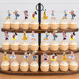 May include: A three-tiered wooden cupcake stand holding twenty-four cupcakes topped with colorful sprinkles and paper cutouts of various cartoon characters including Mickey Mouse, Minnie Mouse, Donald Duck, Daisy Duck, Goofy, and Pluto.
