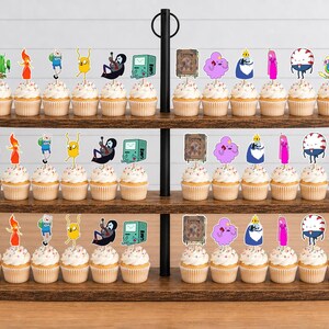 K&ouml;nnte beinhalten: Ein dreist&ouml;ckiger Cupcake-St&auml;nder aus Holz mit zahlreichen Cupcakes, die mit bunten Adventure Time-Figuren aus Papier verziert sind, darunter Finn, Jake, BMO und Prinzessin Bubblegum, angeordnet in Reihen auf jedem ovalen Regalboden.