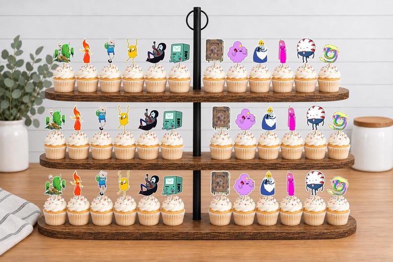 K&ouml;nnte beinhalten: Ein dreist&ouml;ckiger Cupcake-St&auml;nder aus Holz mit zahlreichen Cupcakes, die mit bunten Adventure Time-Figuren aus Papier verziert sind, darunter Finn, Jake, BMO und Prinzessin Bubblegum, angeordnet in Reihen auf jedem ovalen Regalboden.