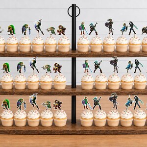 Op de afbeelding: Een houten cupcake-standaard met drie niveaus met talloze cupcakes, elk bekroond met kleine, kleurrijke papieren uitsparingen van diverse videogamepersonages in actiehoudingen, waaronder figuren met zwaarden, schilden en bogen op elke plank.