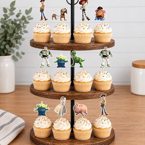 May include: A three-tiered wooden cupcake stand holding twelve cupcakes topped with white frosting, colourful sprinkles, and various character-themed paper toppers including a cowboy, a space ranger, a green alien, a dinosaur, and a pig.