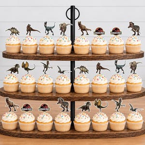 Pu&ograve; includere: Un'alzata per cupcake in legno a tre livelli contenente ventiquattro cupcake guarniti con zuccherini colorati e vari topper di carta a tema dinosauri, inclusi loghi circolari di Jurassic Park, disposti su una superficie in legno marrone.