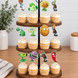 May include: A three-tiered wooden cupcake stand holding twelve cupcakes topped with white frosting and colorful sprinkles. Each cupcake features a decorative paper topper depicting various characters from the Plants vs. Zombies video game.