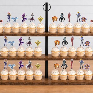 K&ouml;nnte beinhalten: Ein dreist&ouml;ckiger Cupcake-St&auml;nder aus Holz mit zahlreichen Cupcakes, die mit Yu-Gi-Oh!-Papieraufsteckern verziert sind. Die Aufstecker zeigen verschiedene Anime-Figuren, Monster, das Millennium-Puzzle und das rote Yu-Gi-Oh!-Logo auf wei&szlig;em Frosting.