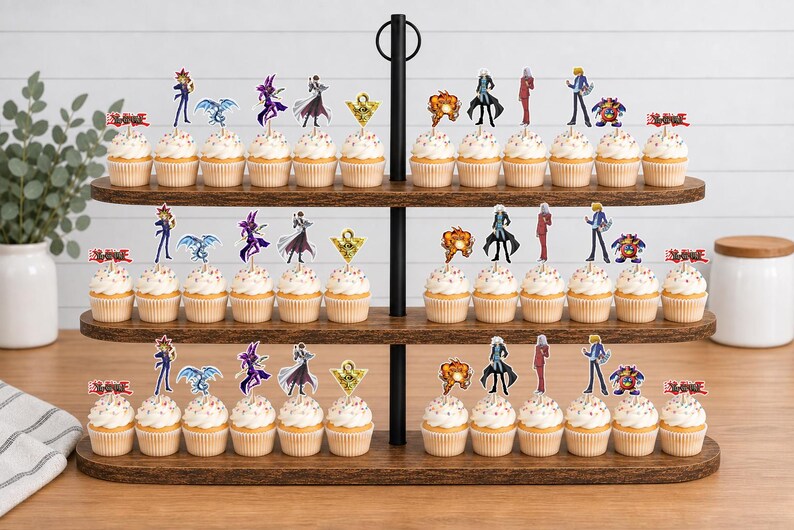 K&ouml;nnte beinhalten: Ein dreist&ouml;ckiger Cupcake-St&auml;nder aus Holz mit zahlreichen Cupcakes, die mit Yu-Gi-Oh!-Papieraufsteckern verziert sind. Die Aufstecker zeigen verschiedene Anime-Figuren, Monster, das Millennium-Puzzle und das rote Yu-Gi-Oh!-Logo auf wei&szlig;em Frosting.