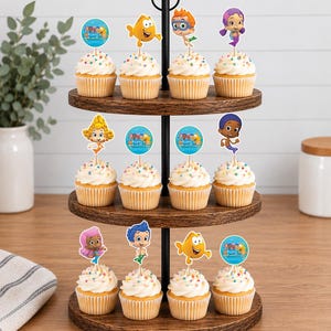 May include: A three-tiered wooden cupcake stand holding twelve cupcakes topped with white frosting, colorful sprinkles, and paper toppers featuring Bubble Guppies characters and logos. The stand has a black metal central handle and frame.