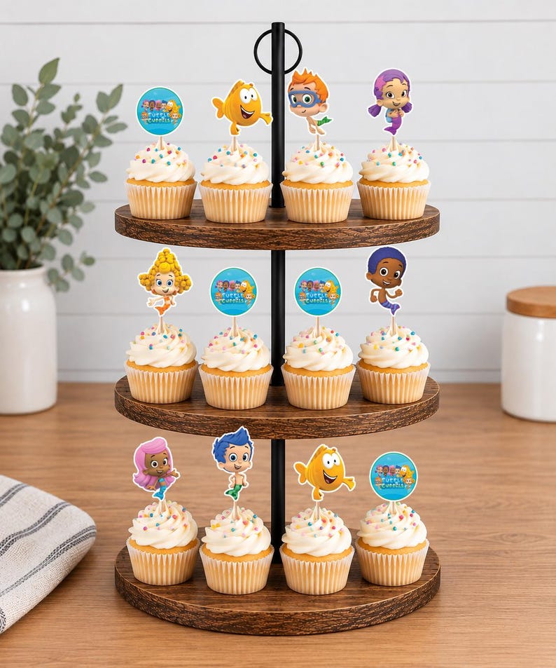 May include: A three-tiered wooden cupcake stand holding twelve cupcakes topped with white frosting, colorful sprinkles, and paper toppers featuring Bubble Guppies characters and logos. The stand has a black metal central handle and frame.