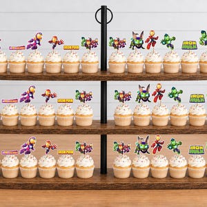 May include: A three-tiered wooden cupcake stand holding numerous cupcakes topped with colorful paper cutouts of various superhero characters, including Iron Man and Hulk, with some featuring text labels like Iron Man and Iron Hulk.