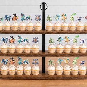 K&ouml;nnte beinhalten: Ein dreist&ouml;ckiger Cupcake-St&auml;nder aus dunklem Holz mit zahlreichen Cupcakes, die mit bunten Papieraufstellern verschiedener animierter Kreaturen und dem Text Pok&eacute;mon Legends verziert sind. Der St&auml;nder hat eine zentrale schwarze Metallstange.