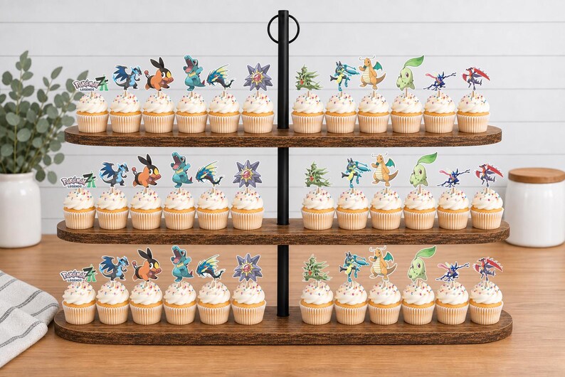 K&ouml;nnte beinhalten: Ein dreist&ouml;ckiger Cupcake-St&auml;nder aus dunklem Holz mit zahlreichen Cupcakes, die mit bunten Papieraufstellern verschiedener animierter Kreaturen und dem Text Pok&eacute;mon Legends verziert sind. Der St&auml;nder hat eine zentrale schwarze Metallstange.