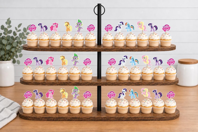 Op de afbeelding: Een houten cupcake-standaard met drie niveaus met talloze cupcakes, bekroond met My Little Pony-papieren toppers. Elke topper bevat een kleurrijke pony- of drakenillustratie en het My Little Pony-logo, gerangschikt in rijen op de donkere houten planken.