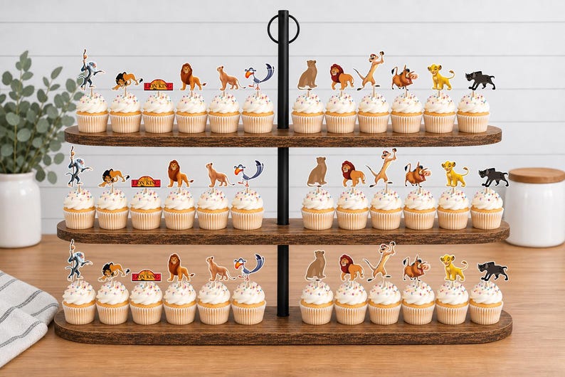 K&ouml;nnte beinhalten: Ein dreist&ouml;ckiger Cupcake-St&auml;nder aus Holz mit zahlreichen Cupcakes, die mit Papieraufstellern von Figuren aus Der K&ouml;nig der L&ouml;wen verziert sind, darunter Simba, Nala, Timon, Pumbaa, Zazu, Rafiki und Mufasa sowie ein rotes Schild mit The Lion King.
