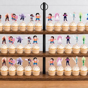 Puede incluir: Un soporte de madera para cupcakes de tres niveles con numerosos cupcakes decorados con recortes de papel de personajes de Steven Universe. Cada cupcake presenta una ilustraci&oacute;n de personaje diferente, incluyendo a Steven, Garnet, Amethyst y Pearl.