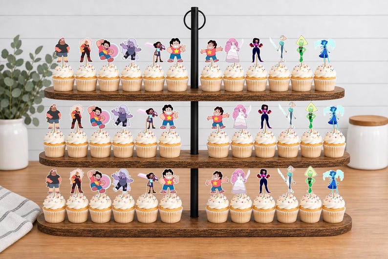 Puede incluir: Un soporte de madera para cupcakes de tres niveles con numerosos cupcakes decorados con recortes de papel de personajes de Steven Universe. Cada cupcake presenta una ilustraci&oacute;n de personaje diferente, incluyendo a Steven, Garnet, Amethyst y Pearl.