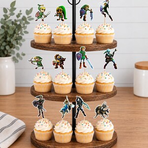 Op de afbeelding: Een houten cupcake-standaard met drie niveaus en twaalf cupcakes, elk voorzien van een toef witte glazuur, kleurrijke hagelslag en een papieren uitsparing van een personage uit de The Legend of Zelda-videogameserie.