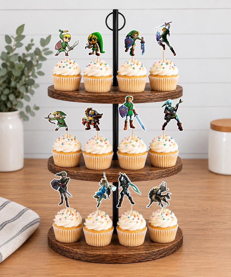 Op de afbeelding: Een houten cupcake-standaard met drie niveaus en twaalf cupcakes, elk voorzien van een toef witte glazuur, kleurrijke hagelslag en een papieren uitsparing van een personage uit de The Legend of Zelda-videogameserie.