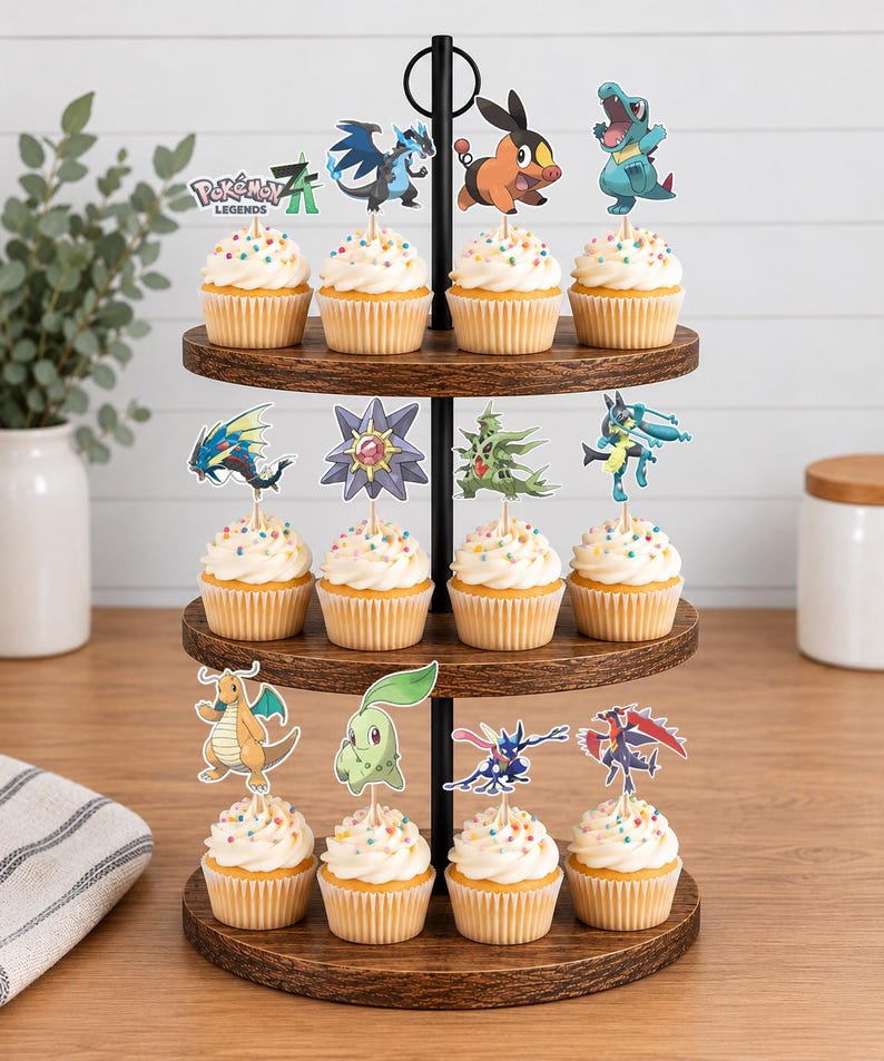 K&ouml;nnte beinhalten: Ein dreist&ouml;ckiger Cupcake-St&auml;nder aus Holz mit zw&ouml;lf Cupcakes, die mit wei&szlig;em Zuckerguss, bunten Streuseln und verschiedenen Pok&eacute;mon-Figuren aus Papier verziert sind, darunter ein Pok&eacute;mon Legends Z-A Logo-Topper.