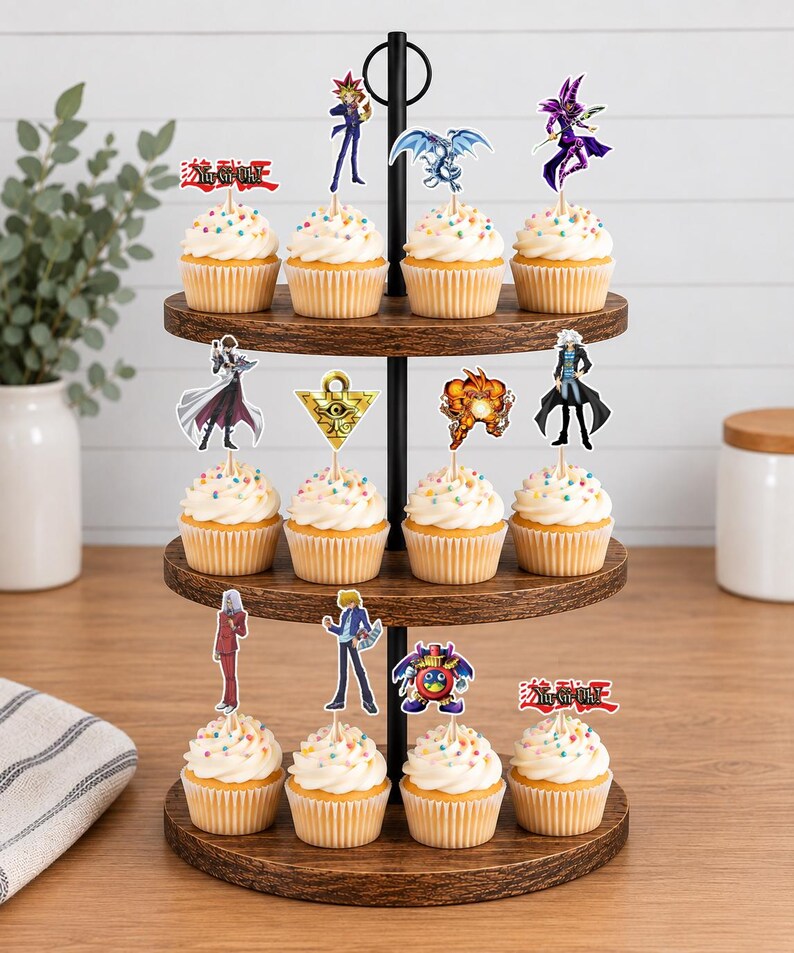 K&ouml;nnte beinhalten: Ein dreist&ouml;ckiger Cupcake-St&auml;nder aus Holz mit zw&ouml;lf Cupcakes, die mit bunten Streuseln und Yu-Gi-Oh!-Papieraufsteckern verziert sind. Die Aufstecker zeigen verschiedene Charaktere, Monster, das Millennium-Puzzle und das rote Logo.