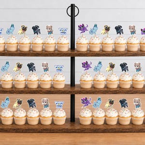 Puede incluir: Un soporte de madera de tres niveles con numerosos cupcakes cubiertos con recortes de papel decorativos. Los adornos presentan personajes de dibujos animados y el texto Puppy Dog Pals. Cada cupcake tiene glaseado blanco y est&aacute; en filas.