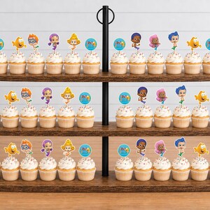 May include: A three-tiered dark wood cupcake stand holding numerous cupcakes topped with colorful Bubble Guppies character paper toppers. Each topper features a cartoon character or a blue circular graphic with text, arranged in rows.