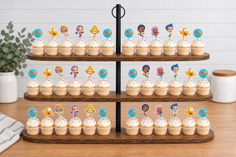 May include: A three-tiered dark wood cupcake stand holding numerous cupcakes topped with colorful Bubble Guppies character paper toppers. Each topper features a cartoon character or a blue circular graphic with text, arranged in rows.