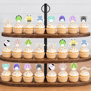 May include: A three-tiered wooden cupcake stand holding twenty-four cupcakes topped with white frosting, colorful sprinkles, and various animal-themed paper toppers. Each topper features a cartoon character, including cows, bees, and koalas.