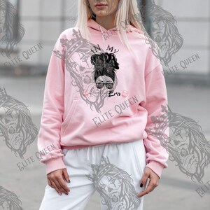 May include: A light pink hoodie with a front graphic of a woman with sunglasses and a messy bun. The text "In My Always Era" is printed on the front. The hoodie has a kangaroo pocket and a drawstring hood.