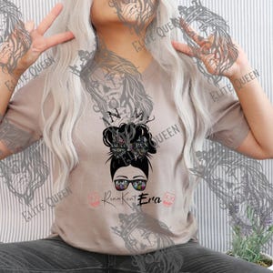 May include: A tan V-neck t-shirt featuring a graphic of a woman's face wearing sunglasses, with a messy bun hairstyle. The text "In My Queen Era" is below the graphic. The shirt has a casual, relaxed fit.