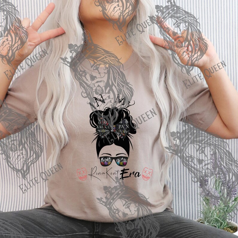May include: A tan V-neck t-shirt featuring a graphic of a woman's face wearing sunglasses, with a messy bun hairstyle. The text "In My Queen Era" is below the graphic. The shirt has a casual, relaxed fit.