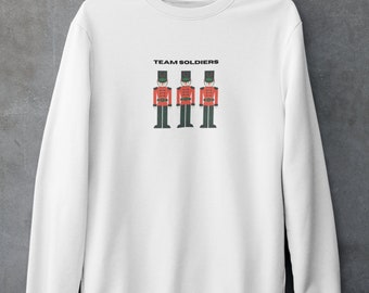 Può includere: Felpa bianca con la scritta "TEAM SOLDIERS" sopra una grafica di tre soldatini con cappotti rossi, pantaloni neri e cappelli verdi. La felpa è appesa a una gruccia color rame contro un muro grigio.