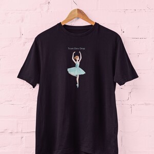 May include: Black t-shirt featuring a ballerina in a light blue tutu. The ballerina is in a graceful pose with arms raised. The text "Team Dew Drop" is above the ballerina. The t-shirt is hanging on a wooden hanger.