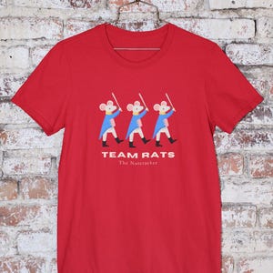 May include: Red t-shirt featuring a graphic of three mice in blue jackets and black boots, holding swords. The text "TEAM RATS" and "The Nutcracker" are printed below the graphic. The shirt is hanging on a metal hanger.