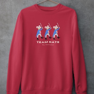 May include: Red sweatshirt with the text "TEAM RATS" and "The Nutcracker." The design features three cartoon mice in blue coats and black boots holding swords. The sweatshirt is hanging on a gold hanger.