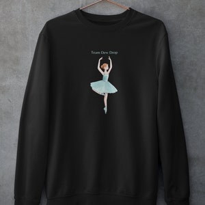 May include: Black sweatshirt featuring a ballerina illustration in a light blue tutu and pointe shoes. The text "Team Dew Drop" is above the ballerina. The sweatshirt is hanging on a copper-colored hanger against a gray wall.