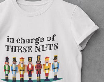 Può includere: T-shirt bianca con la scritta nera "in charge of THESE NUTS". Sotto, sette figure colorate di schiaccianoci. La maglietta è appesa a un appendiabiti di legno.