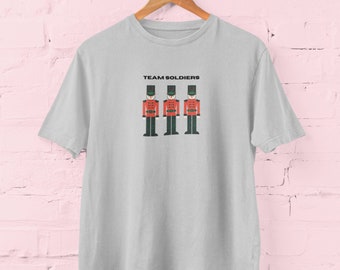 Può includere: Una maglietta grigio chiaro con la scritta "TEAM SOLDIERS" sopra una grafica di tre soldatini. I soldati indossano cappotti rossi, cappelli neri e pantaloni verdi. La maglietta è appesa a una gruccia di legno.