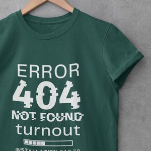 May include: A dark green t-shirt with the text "ERROR 404 NOT FOUND turnout INSTALLATION FAILED" in white. The design has a glitch effect on the numbers. The shirt is hanging on a wooden hanger.